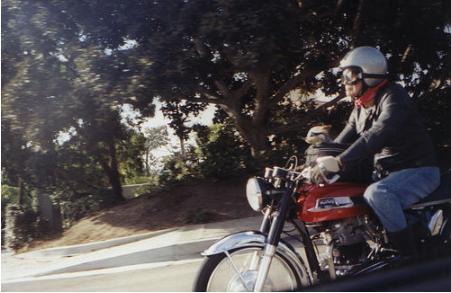 A person wearing a helmet and riding a motorcycle. A man riding a motorcycle with a small dog propped on the fuel tank.