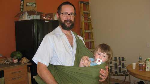 1) What should we describe? What s in this image? What do people describe? A bearded man is holding a child in a sling. A bearded man stands while holding a small child in a green sheet.